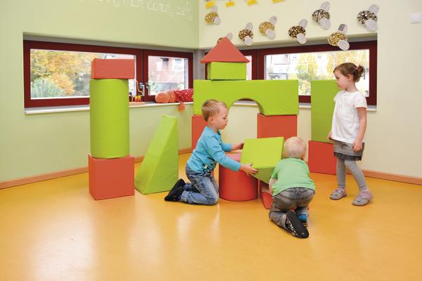 Kinder spielen mit großen bunten Schaumstoffbausteinen in einem hellen Spielzimmer.