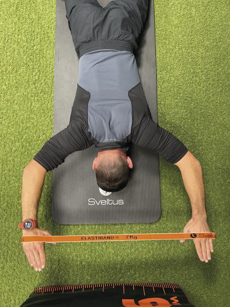 Mann macht Schulterübung mit elastischem Widerstandsband auf Fitnessmatte auf grünem Boden.