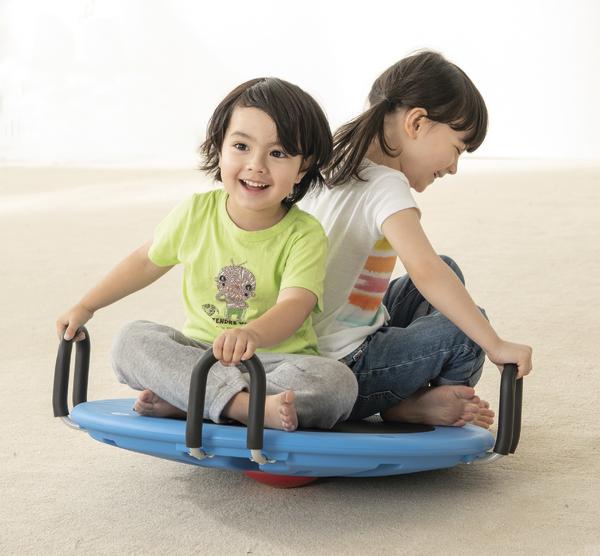 Zwei Kinder sitzen lachend auf einem blauen Wippbrett mit Griffen auf hellem Boden.
