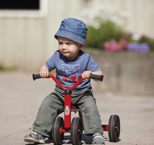 Kleinkind mit blauem Hut fährt rotes Dreirad auf sonnigem Gehweg draußen.