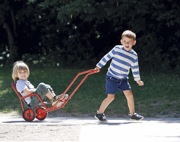 Kind zieht lachend ein anderes Kind im roten Bollerwagen im sonnigen Park spazieren.