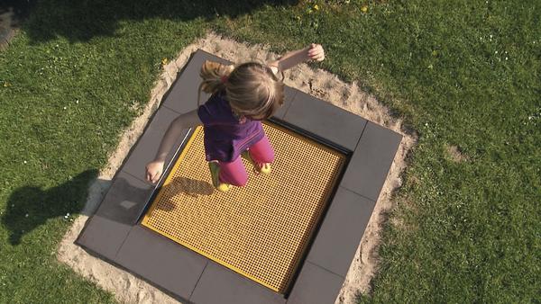 Kind springt auf eingebautem Trampolin im Garten, umgeben von Gras und Sand.