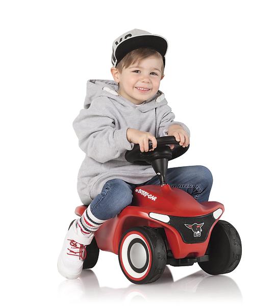 Kind sitzt lachend auf rotem Rutschauto mit schwarzem Lenkrad und grauer Kapuzenjacke.