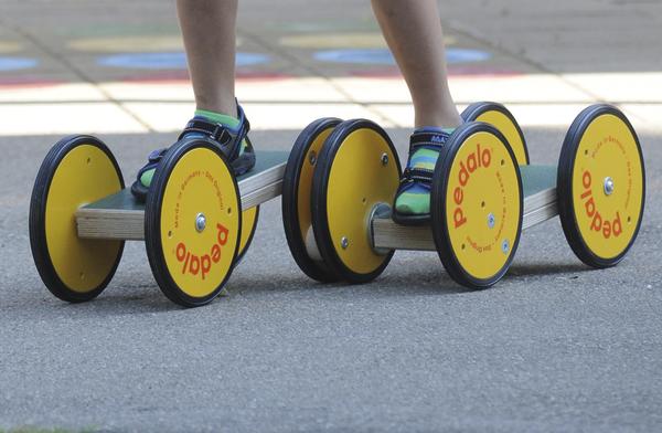 Kind steht auf Pedalo-Brettern mit gelben Rädern auf Asphalt, balanciert.