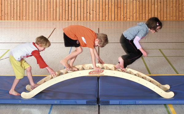 Kinder balancieren auf einem gebogenen Holz-Balancebalken in einer Turnhalle.