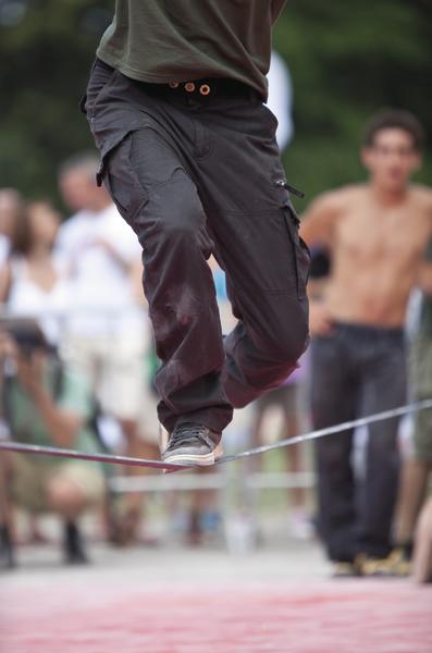 Mann balanciert auf Slackline im Freien vor einer Menschenmenge bei sonnigem Wetter.