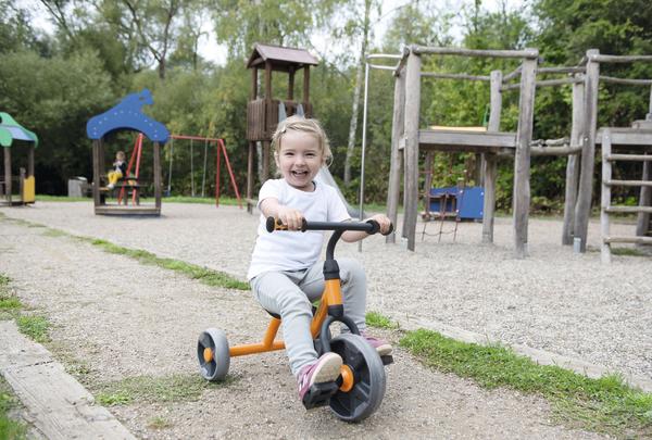 Kind auf Dreirad fährt lachend auf Spielplatz mit Klettergerüst und Rutsche im Hintergrund.