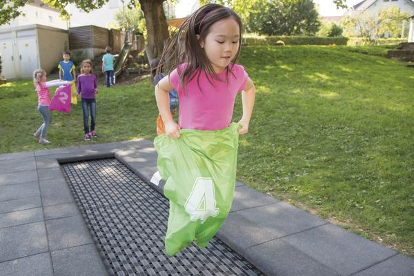 Kind beim Sackhüpfen auf Spielplatz, andere Kinder warten im Hintergrund.