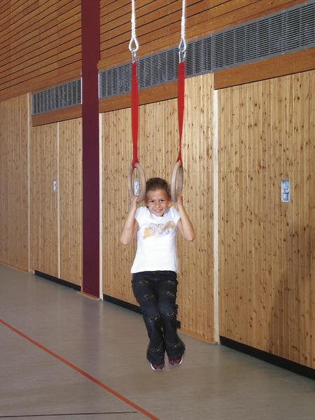 Mädchen schwingt an Turnringen in einer Sporthalle mit Holzverkleidung.