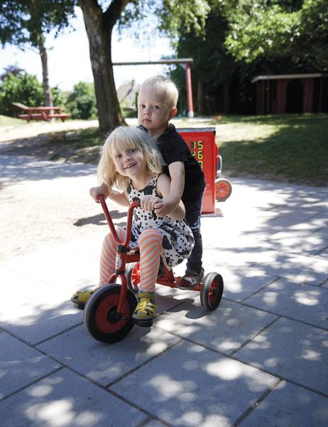Zwei Kinder fahren gemeinsam auf einem roten Dreirad im sonnigen Park.