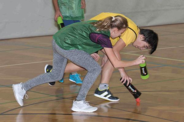 Kinder spielen in der Turnhalle ein Fangspiel mit bunten Flaschen und Bällen auf dem Boden.