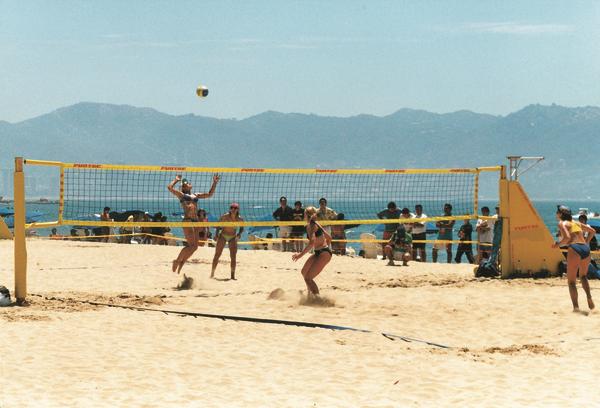 Strandvolleyballspiel mit Spielern und Zuschauern vor Bergkulisse am sonnigen Strand.