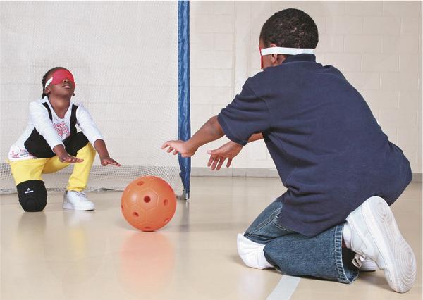 Zwei Kinder mit Augenbinden spielen mit einem taktilen Ball in einer Sporthalle.