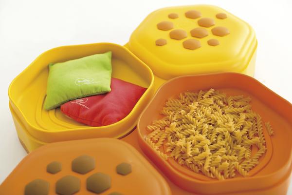 Gelbe und orangefarbene Kunststoffbehälter mit Nudeln und bunten Stoffbeuteln, auf weißem Hintergrund.