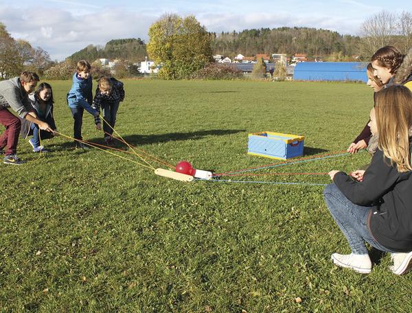Kinder spielen im Freien ein Teamspiel mit Seilen und einem Ball auf einer Wiese.
