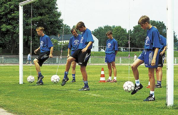 Jugendliche trainieren Fußballtechnik mit Bällen an Schnüren auf grünem Sportplatz.