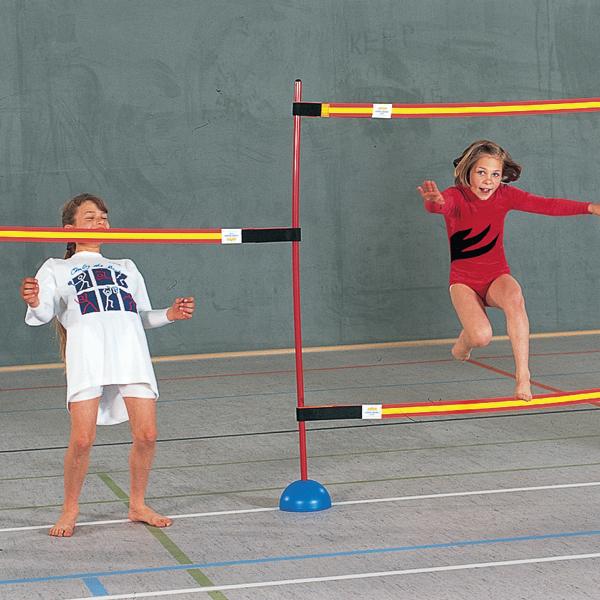 Ein Mädchen in roter Sportkleidung springt in einer Turnhalle über ein elastisches Band, während ein anderes Kind daneben steht und zuschaut.