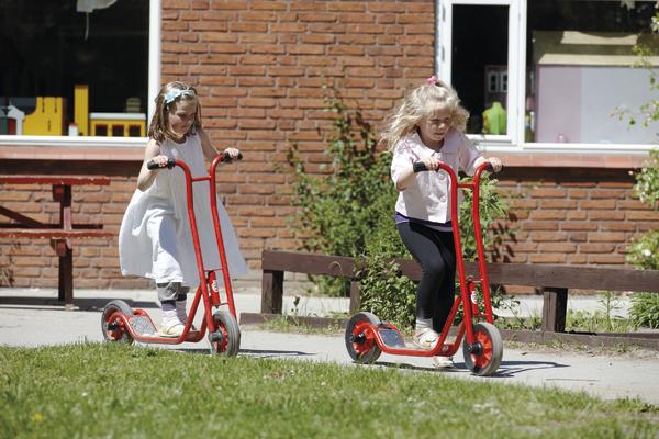 Zwei Mädchen fahren mit roten Rollern auf einem Gehweg vor einem Backsteingebäude.