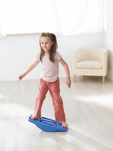 Mädchen balanciert barfuß auf blauem Wackelbrett im hellen Wohnzimmer mit Sessel.
