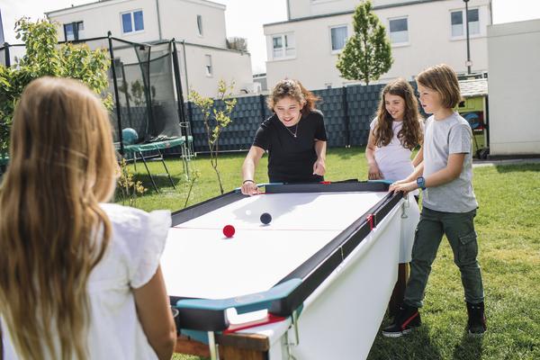 Kinder spielen draußen auf einem Airhockey-Tisch im Garten bei sonnigem Wetter.