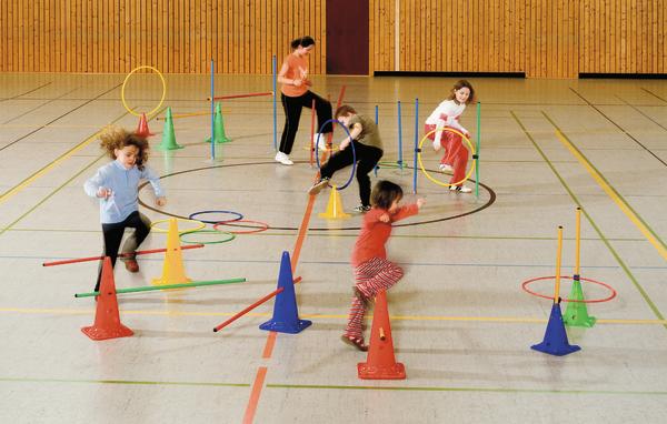 Kinder spielen in einer Turnhalle an einem bunten Hindernisparcours mit Kegeln und Reifen.