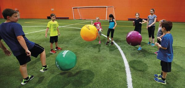 Kinder spielen mit bunten Bällen auf grünem Kunstrasen in einer Halle mit orangefarbener Wand.