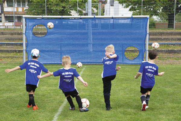 Vier Kinder in blauen Trikots schießen Bälle auf ein Fußballtor mit Zielscheiben auf dem Rasen.