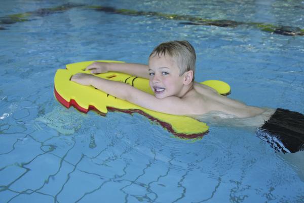 Junge schwimmt im Pool mit gelbem Schwimmbrett und lächelt in die Kamera.
