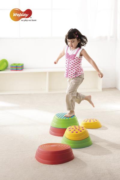 Kind spielt barfuß auf bunten Balancier-Steinen in hellem Raum, Weplay-Logo sichtbar.