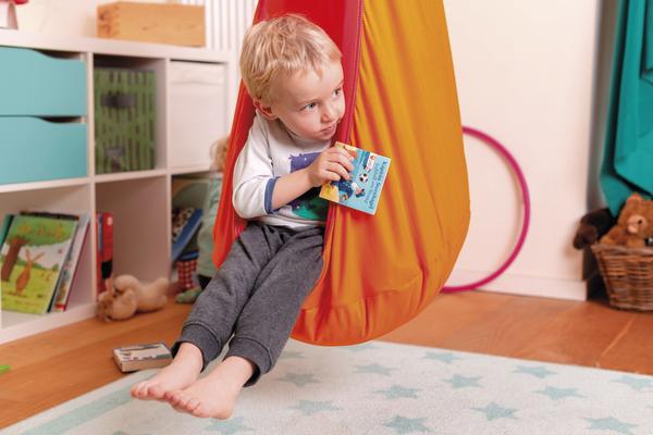 Kind sitzt in orangener Hängeschaukel und liest Kinderbuch im gemütlichen Spielzimmer.