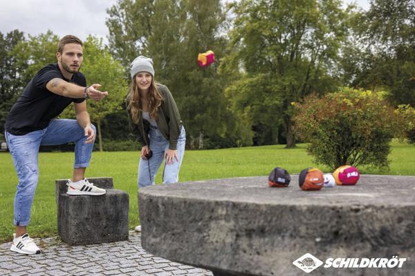 Junge Erwachsene spielen Dosenwerfen im Park auf einer Steinplatte bei grünem Wetter.