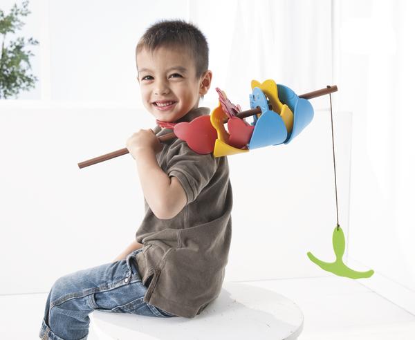 Junge sitzt lächelnd mit selbstgebasteltem Angelspiel aus bunten Papierfischen auf weißem Hintergrund.