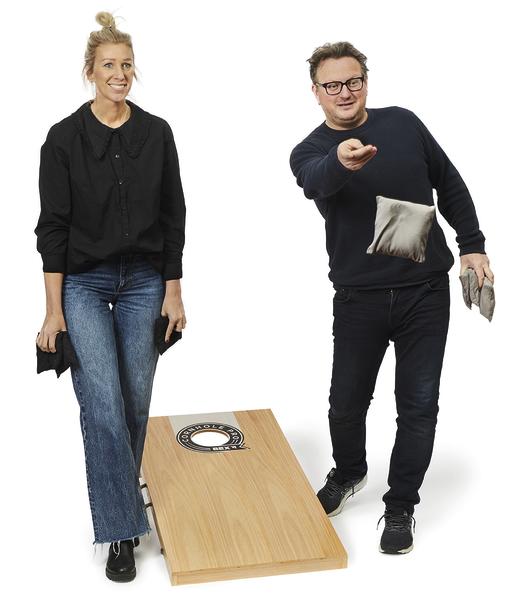 Ein Mann wirft ein Wurfsäckchen auf ein Holz-Spielbrett mit Loch, während eine Frau daneben steht und weitere Säckchen hält.