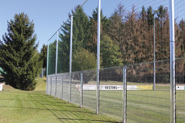 Drahtzaun mit Metallpfosten umgibt grünes Fußballfeld vor Nadelbäumen im Freien.