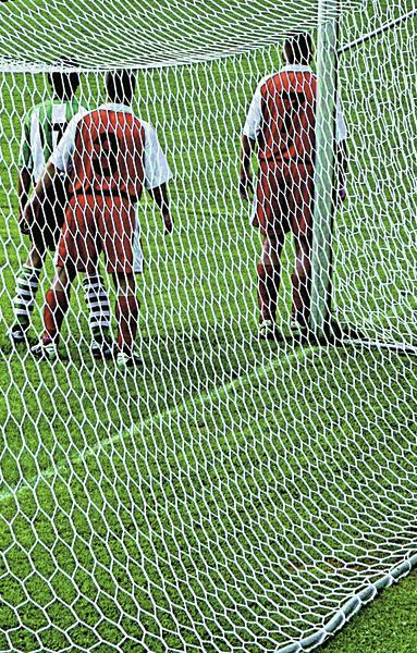 Drei Fußballspieler stehen im Torraum, gesehen durch das Netz des Fußballtors auf grünem Rasen.