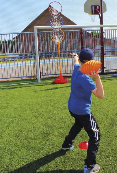 Kind wirft Ball auf Korb im Freien auf Sportplatz mit grünem Kunstrasen.