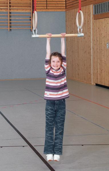 Kind hängt lachend an Turnringen in einer Sporthalle, trägt gestreiftes Shirt und Jeans.