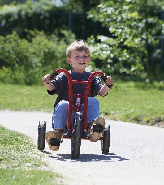 Kind fährt fröhlich auf rotem Dreirad auf einem Weg im Park bei Sonnenschein.