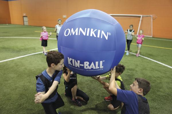 Kinder spielen Kin-Ball mit großem blauen Ball in einer Sporthalle auf Kunstrasen.