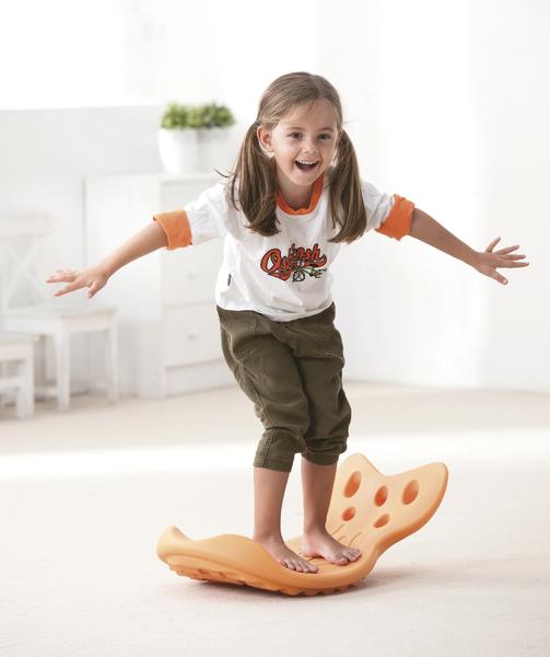 Mädchen balanciert lachend auf orangefarbenem Wackelbrett in hellem Raum mit Pflanzen.