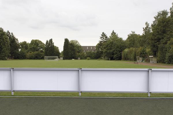 Leerer Fußballplatz mit Toren und Werbebanden vor Bäumen und Gebäuden bei bewölktem Himmel.