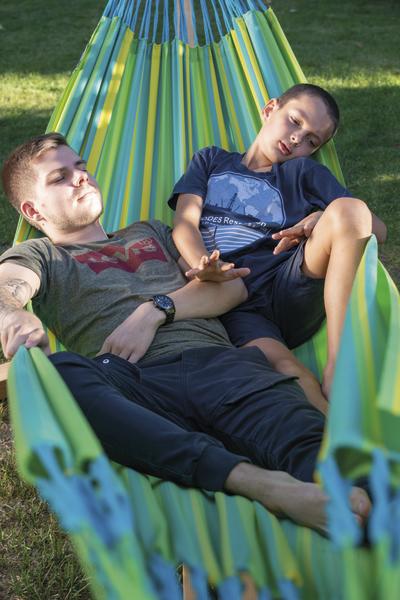 Junger Mann und Junge entspannen gemeinsam in bunter Hängematte im Garten bei Sonnenschein.