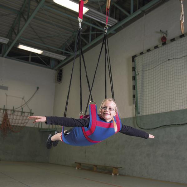 Mädchen in blauer Schaukel mit roten Gurten schwebt in Turnhalle mit ausgestreckten Armen.