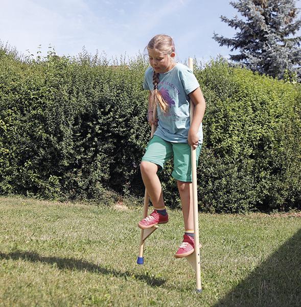 Mädchen übt Stelzenlaufen im Garten an sonnigem Tag vor grüner Hecke.