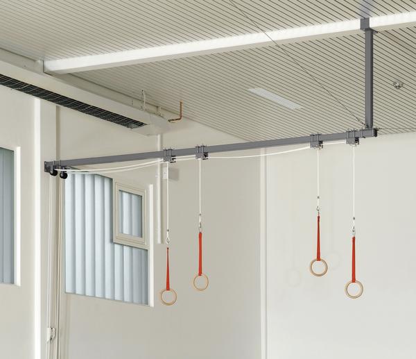 Turnringe mit roten Bändern hängen an einer Deckenhalterung in einem hellen Fitnessraum.