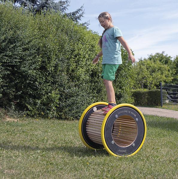 Kind balanciert auf rundem Holzspielgerät im Garten bei Sonnenschein.