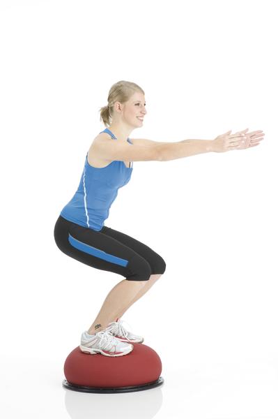 Frau macht Kniebeuge auf rotem Balance-Board in Sportkleidung vor weißem Hintergrund.