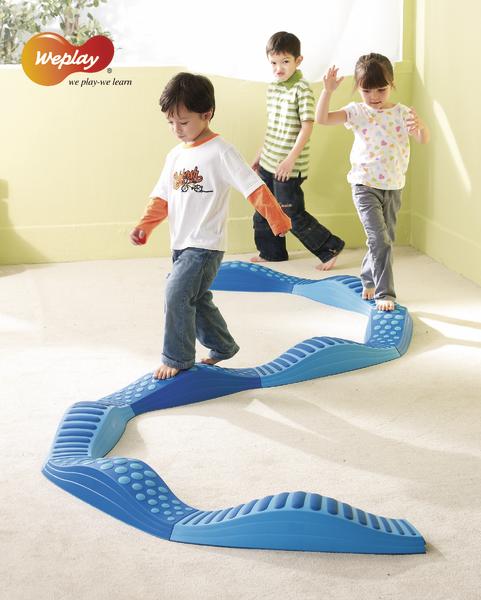 Drei Kinder balancieren auf einem blauen, wellenförmigen Balancierparcours in einem hellen Raum.