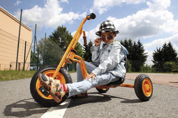 Kind mit Sonnenbrille und Hut fährt orange Dreirad auf Asphalt bei Sonnenschein.