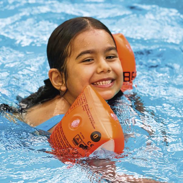 Kind mit orangen Schwimmflügeln lächelt im Schwimmbecken, Wasser umgibt sie.